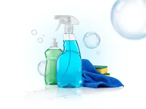 Frasco de detergente verde e borrifador de limpador azul, ao lado de um pano de microfibra e esponjas
